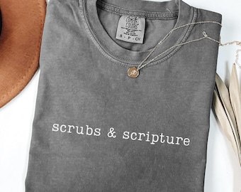 May include: Pepper-colored t-shirt with the words "scrubs & scripture" in white. A silver necklace with a round pendant rests on the shirt. The shirt is folded and displayed with a brown hat and dried leaves.
