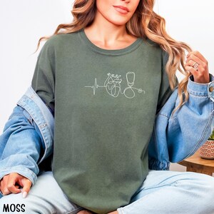 May include: Moss green crew neck sweatshirt featuring a white line drawing of a heart, stethoscope, and heartbeat. The word "MOSS" is printed in white at the bottom left. The model is wearing a denim jacket.