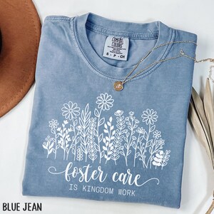 May include: Blue jean Comfort Colors t-shirt with a white floral design and the text "foster care is kingdom work." A gold necklace with a pendant is draped over the shirt. The shirt is folded.