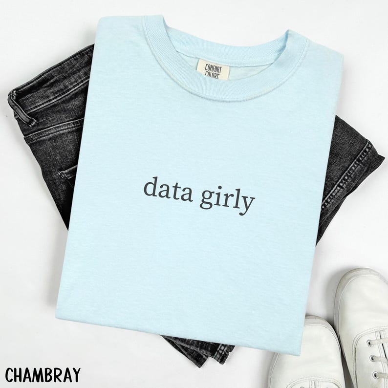 May include: A light blue t-shirt with the text "data girly" in black. The shirt is folded and placed on top of black denim jeans. White sneakers are visible at the bottom right. The shirt has a tag that says "Comfort Colors".