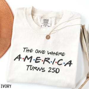 K&ouml;nnte beinhalten: Elfenbeinfarbenes T-Shirt mit dem schwarzen Text "THE ONE WHERE AMERICA TURNS 250", wobei das Wort "AMERICA" in den Farben der amerikanischen Flagge gehalten ist. Eine goldene Halskette mit Anh&auml;nger liegt auf dem Shirt.