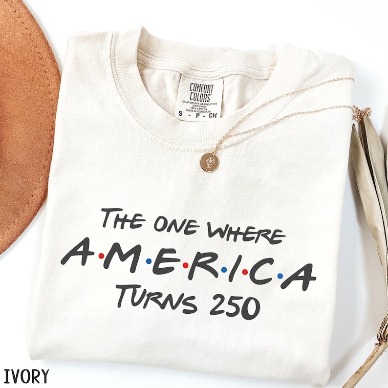 K&ouml;nnte beinhalten: Elfenbeinfarbenes T-Shirt mit dem schwarzen Text "THE ONE WHERE AMERICA TURNS 250", wobei das Wort "AMERICA" in den Farben der amerikanischen Flagge gehalten ist. Eine goldene Halskette mit Anh&auml;nger liegt auf dem Shirt.