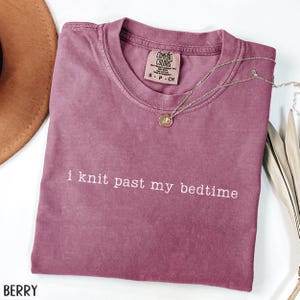 Puede incluir: Camiseta color bayas con el texto blanco "i knit past my bedtime". La camiseta tiene cuello redondo y una peque&ntilde;a etiqueta en el cuello. Un collar dorado con un colgante est&aacute; drapeado sobre la camiseta. Un sombrero marr&oacute;n es visible en la esquina superior izquierda.