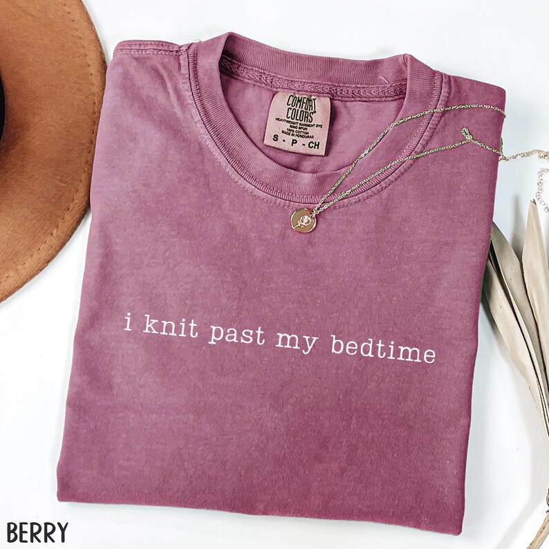 Puede incluir: Camiseta color bayas con el texto blanco "i knit past my bedtime". La camiseta tiene cuello redondo y una peque&ntilde;a etiqueta en el cuello. Un collar dorado con un colgante est&aacute; drapeado sobre la camiseta. Un sombrero marr&oacute;n es visible en la esquina superior izquierda.