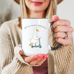 May include: White ceramic mug with a black interior and handle. The mug features a cartoon goose riding a blue scooter, with the text "Off to assess behavior" in black lettering.