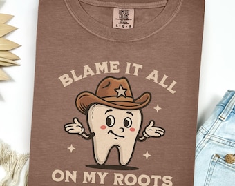 May include: A brown Comfort Colors t-shirt featuring a cartoon tooth wearing a cowboy hat. The text on the shirt reads "BLAME IT ALL ON MY ROOTS". The shirt is folded and next to a pair of blue jeans.