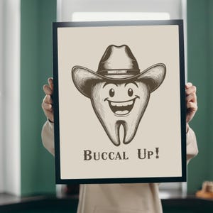 May include: A black framed print with a cartoon tooth wearing a cowboy hat. The tooth is smiling and the text "Buccal Up!" is below the image.