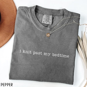 Puede incluir: Una camiseta gris Comfort Colors con el texto "i knit past my bedtime" en blanco. Un collar plateado con un colgante redondo descansa sobre la camiseta. Un sombrero marr&oacute;n es parcialmente visible en la esquina superior izquierda.