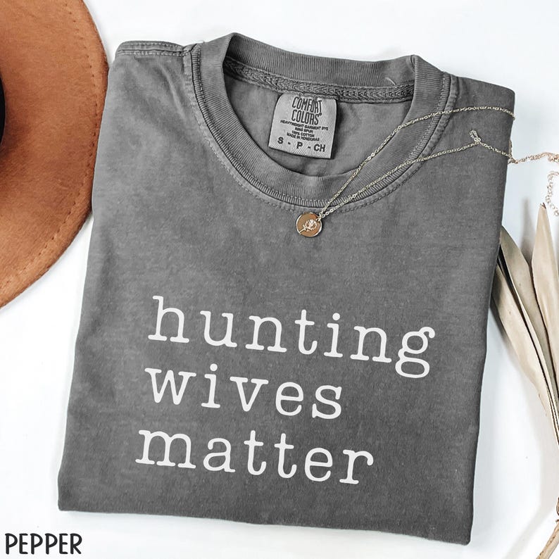K&ouml;nnte beinhalten: Ein graues Comfort Colors T-Shirt mit dem wei&szlig;en Text "hunting wives matter". Eine silberne Halskette mit Anh&auml;nger liegt &uuml;ber dem Shirt. Ein brauner Hut ist oben links zu sehen.