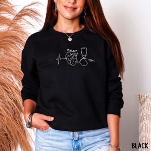 May include: Black sweatshirt with a white line drawing of a heartbeat, heart, and stethoscope. The word "BLACK" is printed in white at the bottom right corner. The sweatshirt is a crew neck style.
