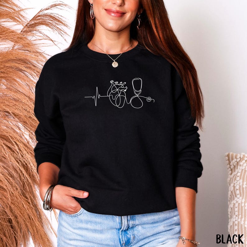 May include: Black sweatshirt with a white line drawing of a heartbeat, heart, and stethoscope. The word "BLACK" is printed in white at the bottom right corner. The sweatshirt is a crew neck style.
