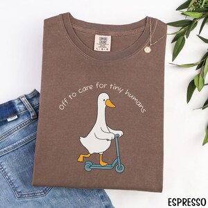 May include: Espresso-colored t-shirt with a cartoon goose riding a blue scooter, and the text "Off to care for tiny humans." The shirt has a Comfort Colors tag and is paired with blue jeans.