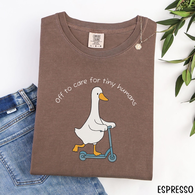 May include: Espresso-colored t-shirt with a cartoon goose riding a blue scooter, and the text "Off to care for tiny humans." The shirt has a Comfort Colors tag and is paired with blue jeans.