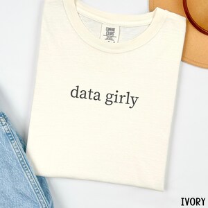 May include: Ivory-colored t-shirt with the phrase "data girly" printed in black. The shirt is made of a soft material and has a classic crew neck design. The shirt is folded and placed on a white surface.