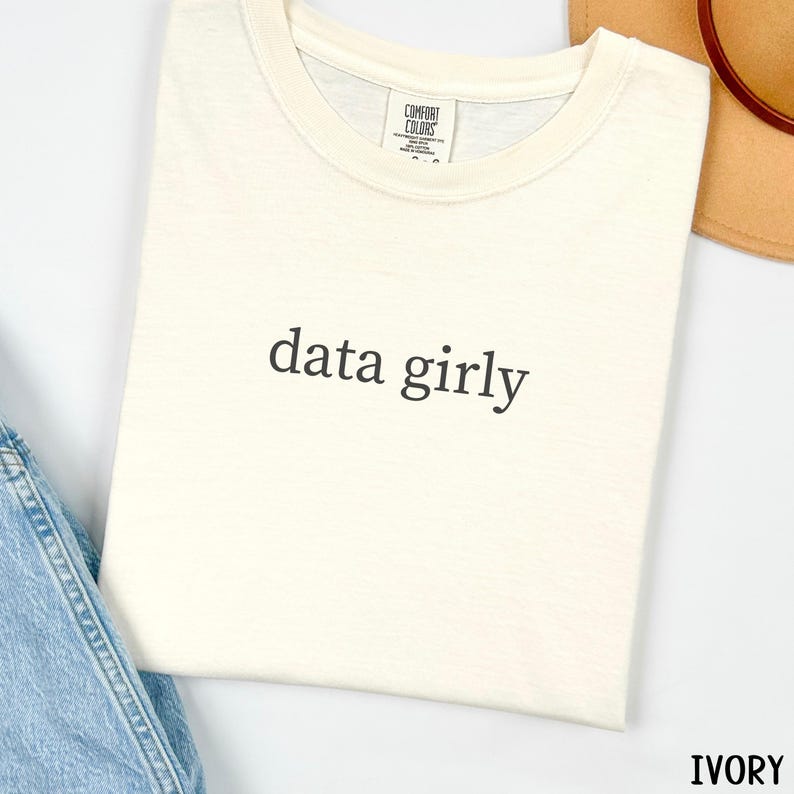 May include: Ivory-colored t-shirt with the phrase "data girly" printed in black. The shirt is made of a soft material and has a classic crew neck design. The shirt is folded and placed on a white surface.