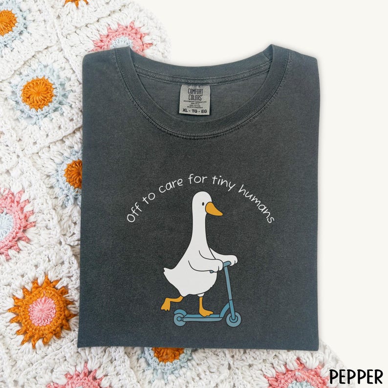 May include: Pepper-colored Comfort Colors t-shirt with a graphic of a white goose riding a blue scooter. The text "off to care for tiny humans" is arched above the goose. The shirt is folded on a white and pastel-colored crocheted blanket.