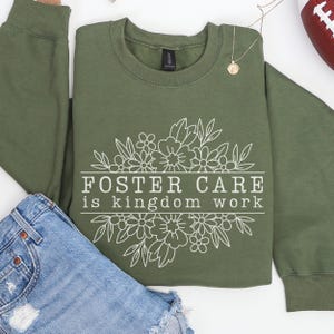 May include: Olive green sweatshirt with the text "FOSTER CARE is KINGDOM WORK" in white, surrounded by a floral design. The sweatshirt is paired with denim shorts and a gold pendant necklace.