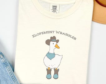 May include: Cream-colored t-shirt featuring a cartoon goose wearing a cowboy hat, neckerchief, and boots. The text "ELOPEMENT WRANGLER" is arched above the goose. The shirt is folded, with a tag visible at the neckline.