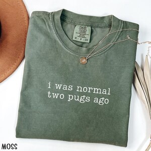 May include: A moss green t-shirt with the slogan "i was normal two pugs ago" in white lettering. The shirt has a crew neck and short sleeves. A gold necklace with a pendant is draped across the shirt.