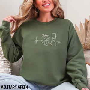 May include: Military green sweatshirt featuring a white line drawing of a heart, stethoscope, and heartbeat rhythm. The design is centered on the chest. The sweatshirt is a solid color and has a crew neck.