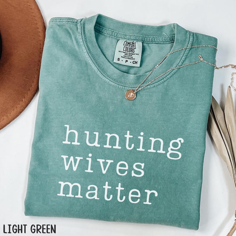 K&ouml;nnte beinhalten: Hellgr&uuml;nes T-Shirt mit dem wei&szlig;en Text "hunting wives matter". Das Shirt hat einen Rundhalsausschnitt und kurze &Auml;rmel. Eine silberne Halskette mit rundem Anh&auml;nger liegt &uuml;ber dem Shirt. Das Shirt besteht aus einem weichen, bequemen Material.