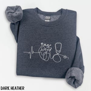 May include: Dark heather gray sweatshirt with a white line drawing of a heart, heartbeat, and stethoscope. The text "DARK HEATHER" is at the bottom of the image. The sweatshirt has a crew neck and cuffed sleeves.