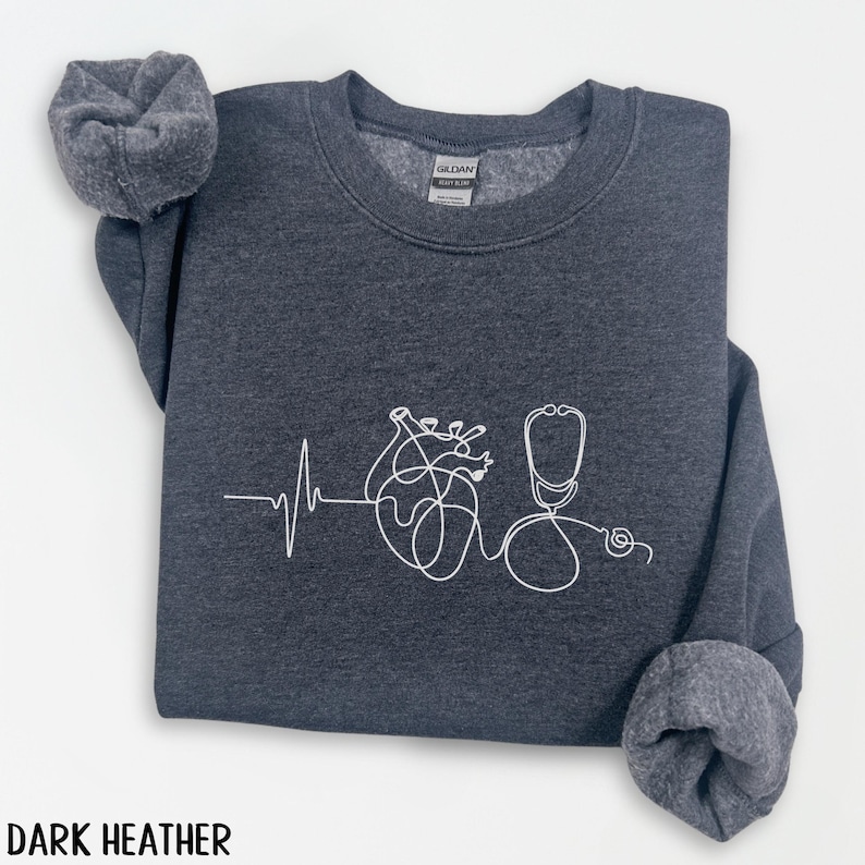 May include: Dark heather gray sweatshirt with a white line drawing of a heart, heartbeat, and stethoscope. The text "DARK HEATHER" is at the bottom of the image. The sweatshirt has a crew neck and cuffed sleeves.