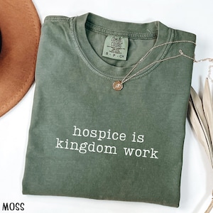 May include: A moss green t-shirt with the text "hospice is kingdom work" in white. The shirt has a crew neck and short sleeves. A gold-colored necklace with a pendant is draped across the shirt. The shirt is folded and has a tag that says "Comfort Colors".