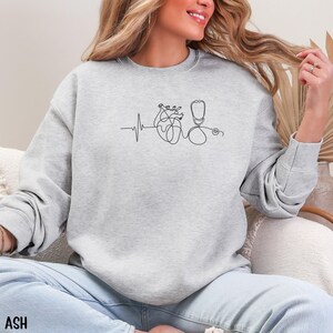 May include: Light gray sweatshirt with a black line drawing of a heart, stethoscope, and heartbeat. The word "ASH" is printed in the bottom left corner. The sweatshirt is a crew neck style.