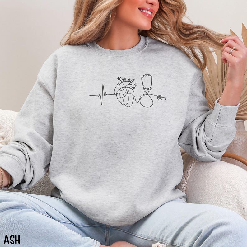 May include: Light gray sweatshirt with a black line drawing of a heart, stethoscope, and heartbeat. The word "ASH" is printed in the bottom left corner. The sweatshirt is a crew neck style.