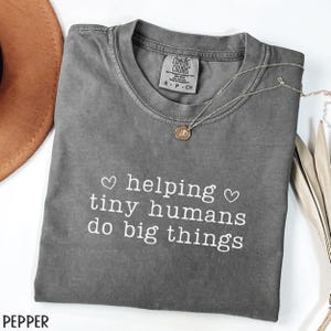 May include: A gray Comfort Colors t-shirt with the text "helping tiny humans do big things" in white. A silver necklace with a pendant is draped across the shirt. The shirt is folded and displayed with a brown hat.