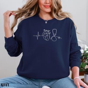 May include: Navy blue sweatshirt featuring a white line drawing of a heartbeat, a heart, and a stethoscope. The word "NAVY" is printed at the bottom of the image. The sweatshirt is a solid color and has a crew neck.