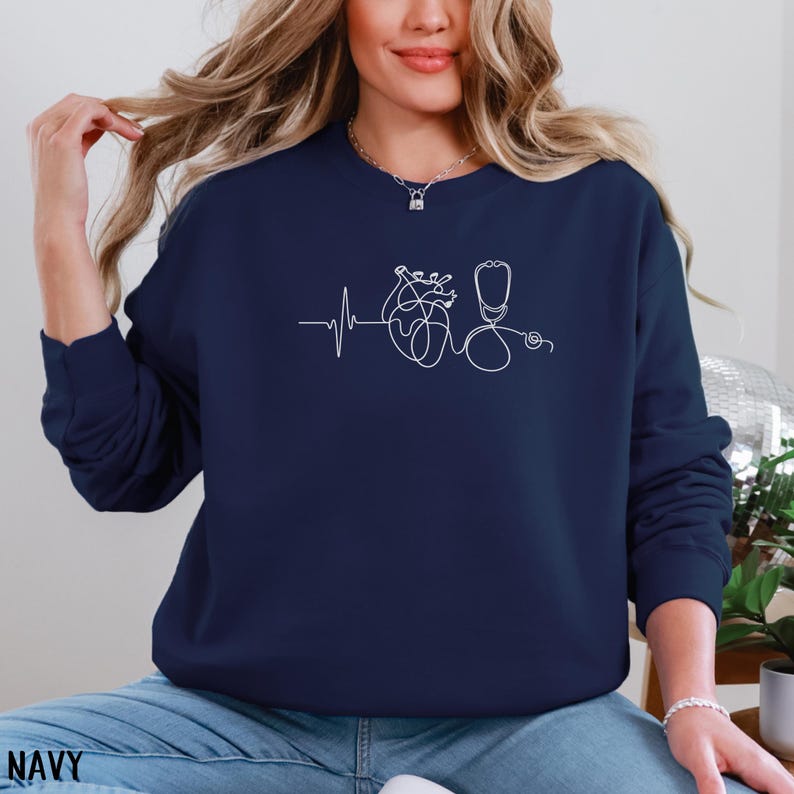 May include: Navy blue sweatshirt featuring a white line drawing of a heartbeat, a heart, and a stethoscope. The word "NAVY" is printed at the bottom of the image. The sweatshirt is a solid color and has a crew neck.