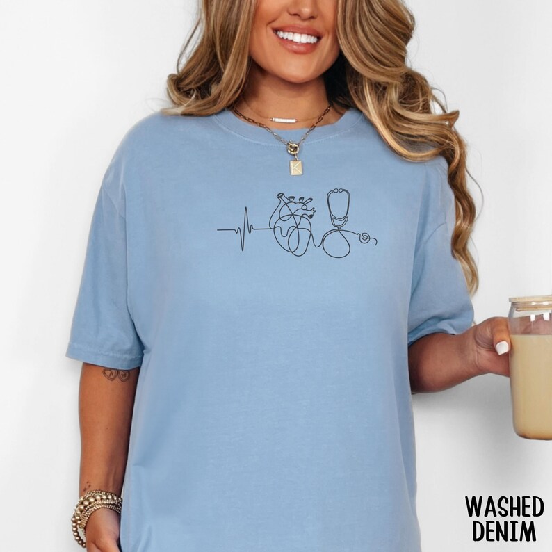 May include: Pale blue t-shirt featuring a black line drawing of a heartbeat, heart, and stethoscope. The shirt has short sleeves and a loose fit. The words "Washed Denim" are printed in the lower right corner.