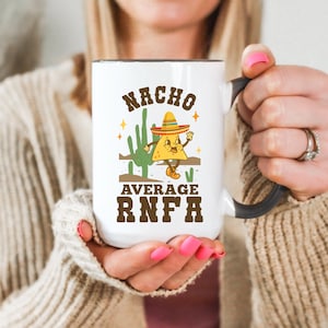 May include: A white ceramic mug with a dark gray handle featuring a graphic of a smiling tortilla chip wearing a sombrero and boots standing next to a green cactus. The text reads NACHO AVERAGE RNFA in brown, stylized, bold, block lettering.