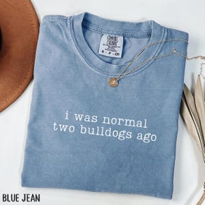 May include: A blue jean-colored Comfort Colors t-shirt with the phrase "i was normal two bulldogs ago" in white lettering. A gold necklace with a pendant rests on the shirt. The shirt is folded and displayed on a white surface.