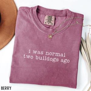 May include: Berry-colored Comfort Colors t-shirt with the text "i was normal two bulldogs ago" in white. A gold necklace with a pendant is draped across the shirt. A brown hat is visible in the upper left corner.