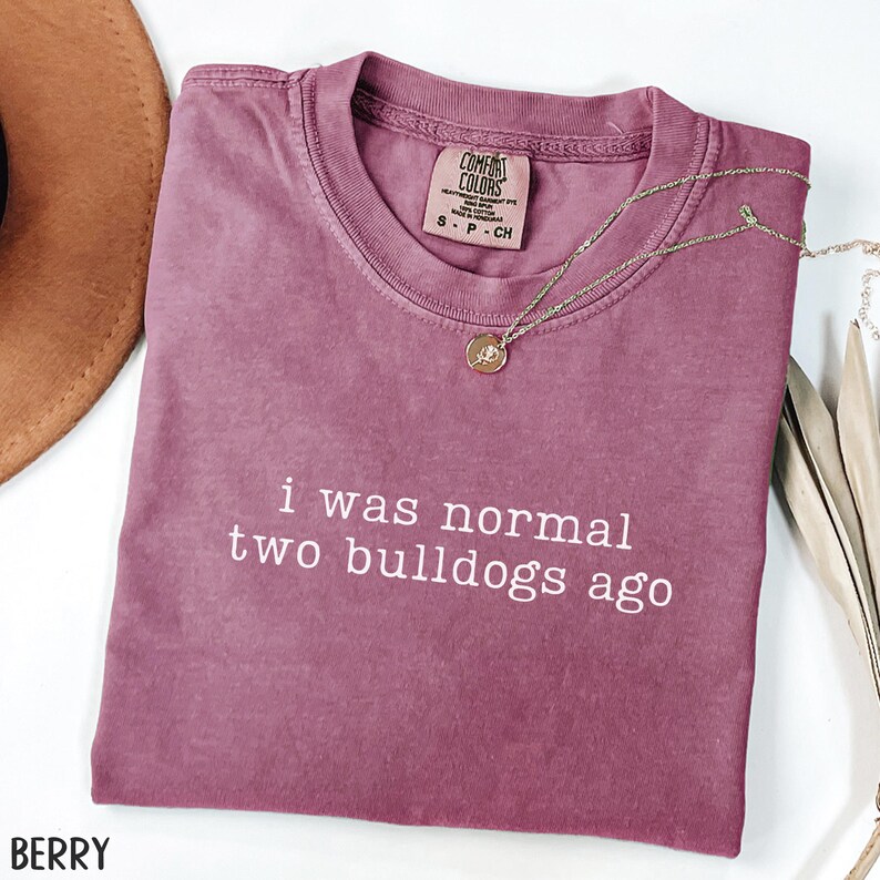 May include: Berry-colored Comfort Colors t-shirt with the text "i was normal two bulldogs ago" in white. A gold necklace with a pendant is draped across the shirt. A brown hat is visible in the upper left corner.