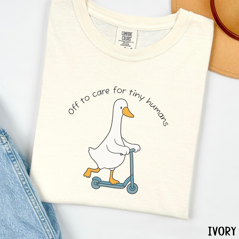 May include: Ivory-colored t-shirt featuring a cartoon goose riding a blue scooter. The text "off to care for tiny humans" is arched above the goose. The shirt has a Comfort Colors tag.