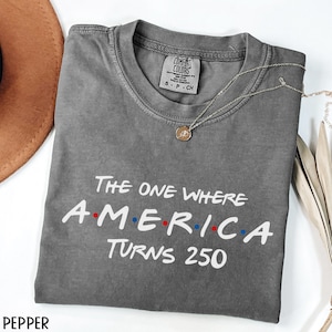 K&ouml;nnte beinhalten: Ein graues Comfort Colors T-Shirt mit dem Text "The One Where America Turns 250" in Wei&szlig;, Rot und Blau. Eine silberne Halskette mit einem runden Anh&auml;nger liegt auf dem T-Shirt. Das T-Shirt ist auf einer wei&szlig;en Oberfl&auml;che gefaltet.