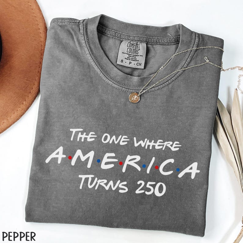 K&ouml;nnte beinhalten: Ein graues Comfort Colors T-Shirt mit dem Text "The One Where America Turns 250" in Wei&szlig;, Rot und Blau. Eine silberne Halskette mit einem runden Anh&auml;nger liegt auf dem T-Shirt. Das T-Shirt ist auf einer wei&szlig;en Oberfl&auml;che gefaltet.