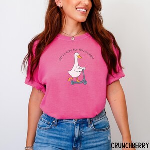 May include: A bright pink t-shirt with the text "off to care for tiny humans" arched above a cartoon goose riding a blue scooter. The goose is white with a yellow beak and legs. The shirt is made of a soft material.