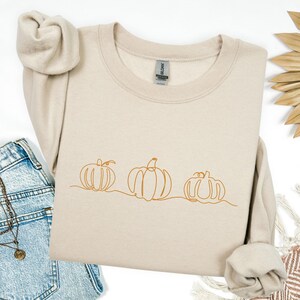 May include: Beige sweatshirt featuring a line drawing of three pumpkins in a row. The pumpkins are drawn in a continuous line with a minimalist style. The sweatshirt is folded, with the sleeves and a portion of the jeans visible.