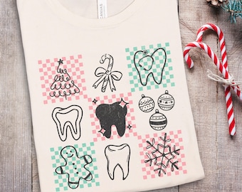 May include: Cream-colored t-shirt featuring a grid of Christmas-themed dental illustrations. Designs include a Christmas tree, candy cane, tooth with a stethoscope, ornaments, a gingerbread man, a sparkling tooth, and a snowflake. The shirt is laid flat on a wooden surface.