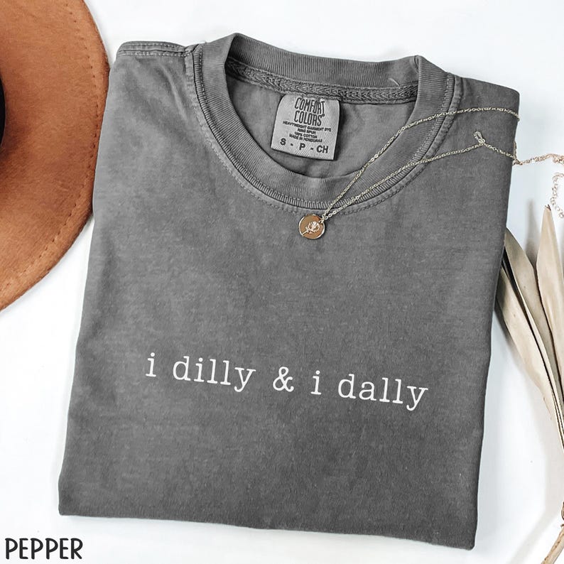 May include: A gray Comfort Colors t-shirt with the phrase "i dilly & i dally" in white. A silver necklace with a small pendant rests on the shirt. The shirt is folded and displayed with a brown hat and decorative leaves.