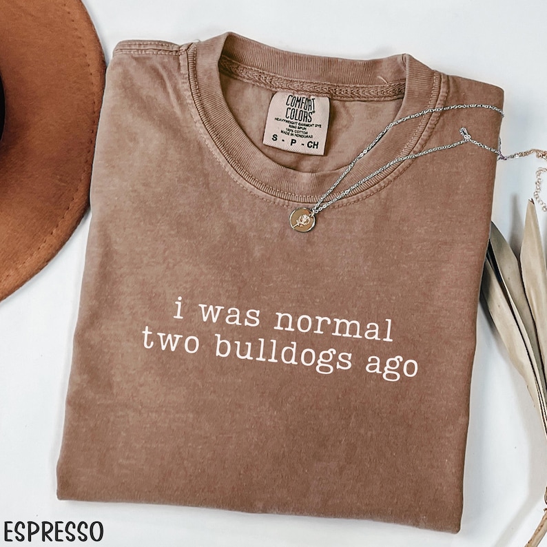 May include: Espresso-colored t-shirt with the text "i was normal two bulldogs ago" in white. A silver necklace with a pendant is draped across the shirt. The shirt is folded and has a Comfort Colors tag.