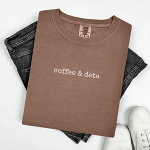 May include: A brown crew neck sweatshirt with the words "coffee & data" in white script. The sweatshirt is folded on top of a pair of dark wash jeans. White sneakers are visible at the bottom of the image.