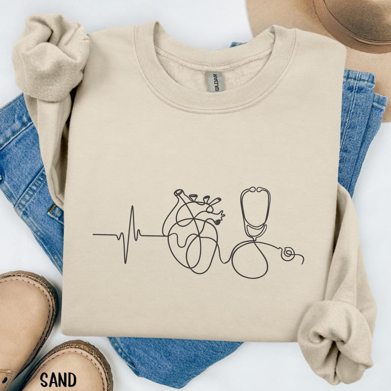 May include: Sand-colored sweatshirt featuring a black line drawing of a heart, a stethoscope, and a heartbeat rhythm. The sweatshirt is made of a soft material and is a crew neck style. The word "SAND" is printed on the bottom of the image.