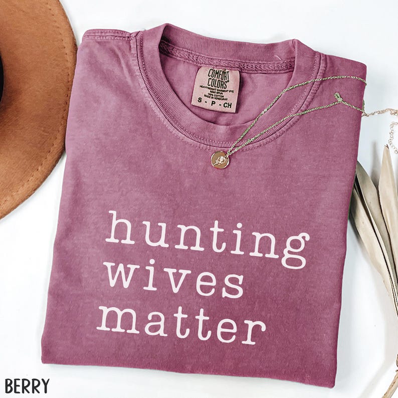 K&ouml;nnte beinhalten: Ein beerenfarbenes T-Shirt mit dem wei&szlig;en Text "hunting wives matter". Eine goldene Halskette mit Anh&auml;nger liegt &uuml;ber dem Shirt. Das Shirt ist gefaltet und hat ein Comfort Colors-Etikett.