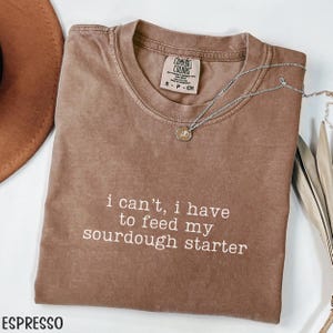 May include: Espresso-colored t-shirt with the text "i can't, i have to feed my sourdough starter" in white. The shirt has a crew neck and short sleeves. A silver necklace with a pendant is draped across the shirt.